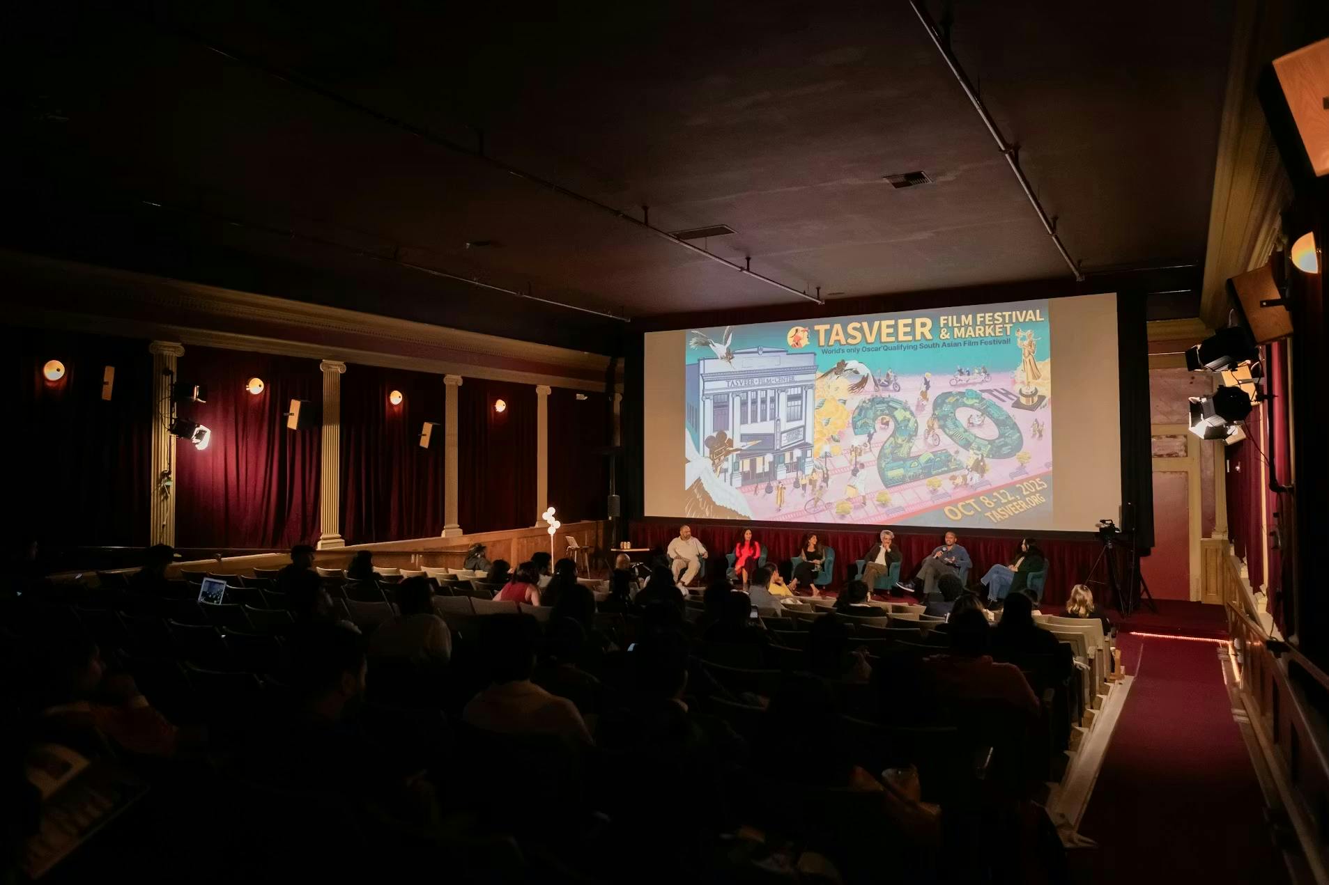 Image of a Tasveer Film Festival Theater displaying Tasveer Film Festival and Market poster. Panel sitting in front of screen and audience facing the panel. 
