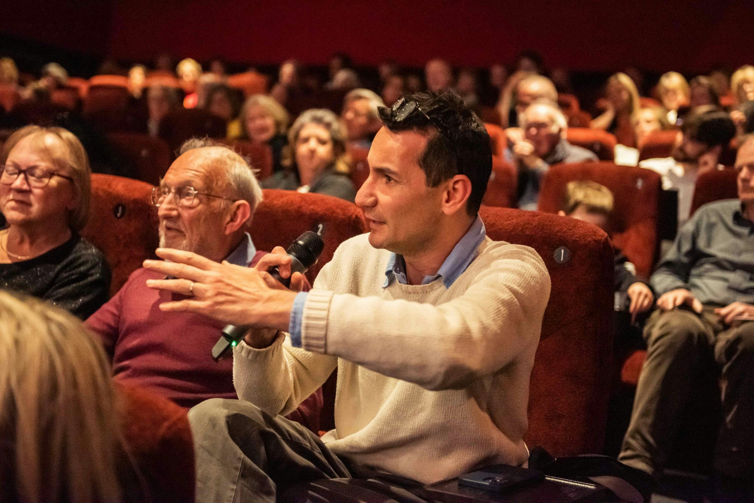 A man, Andrea, holds a microwave asking a question to an on stage guest while sitting in a full audience.