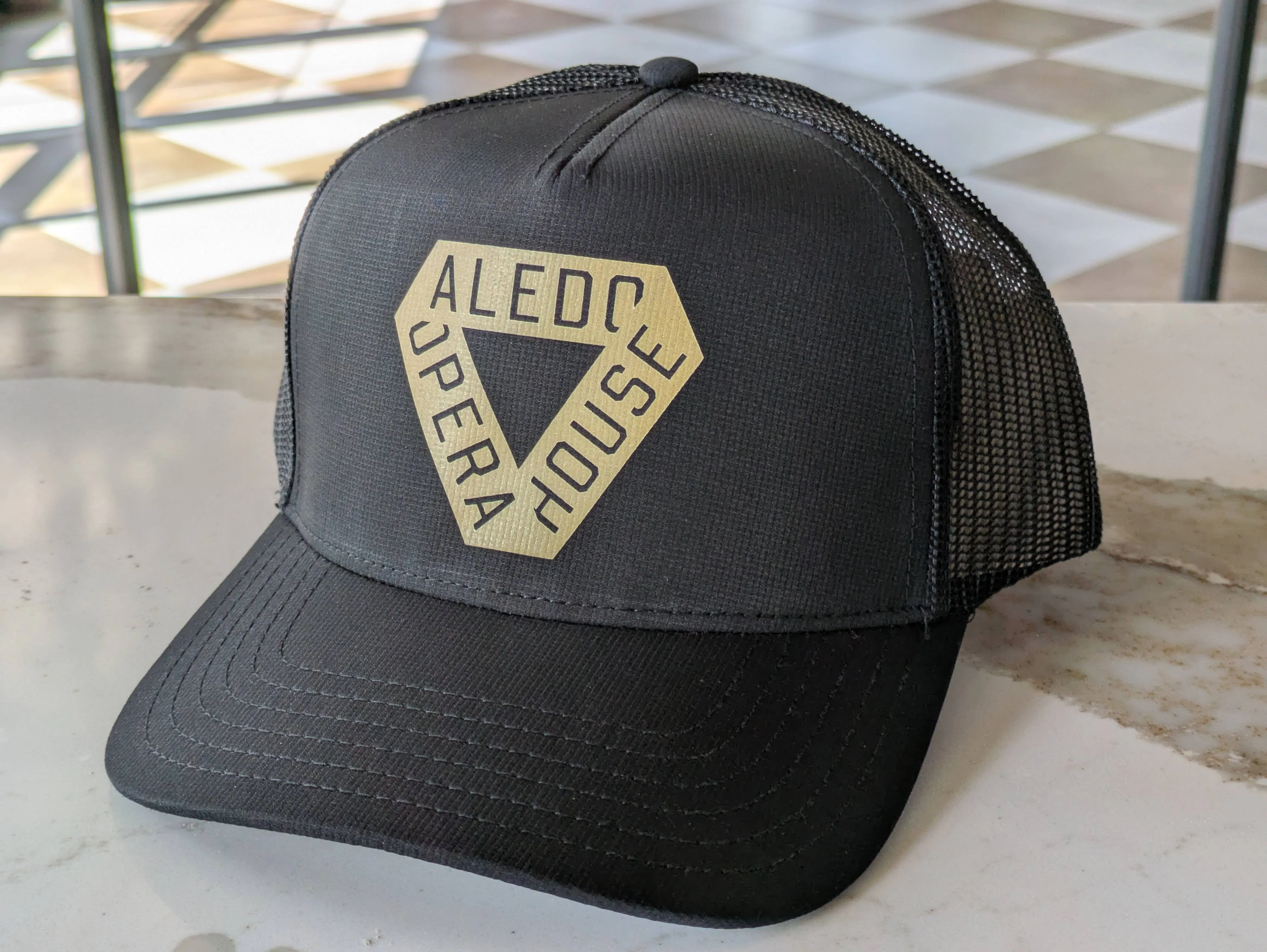 a black trucker hat with a golden triangle delta ribbon and the words Aledo Opera House