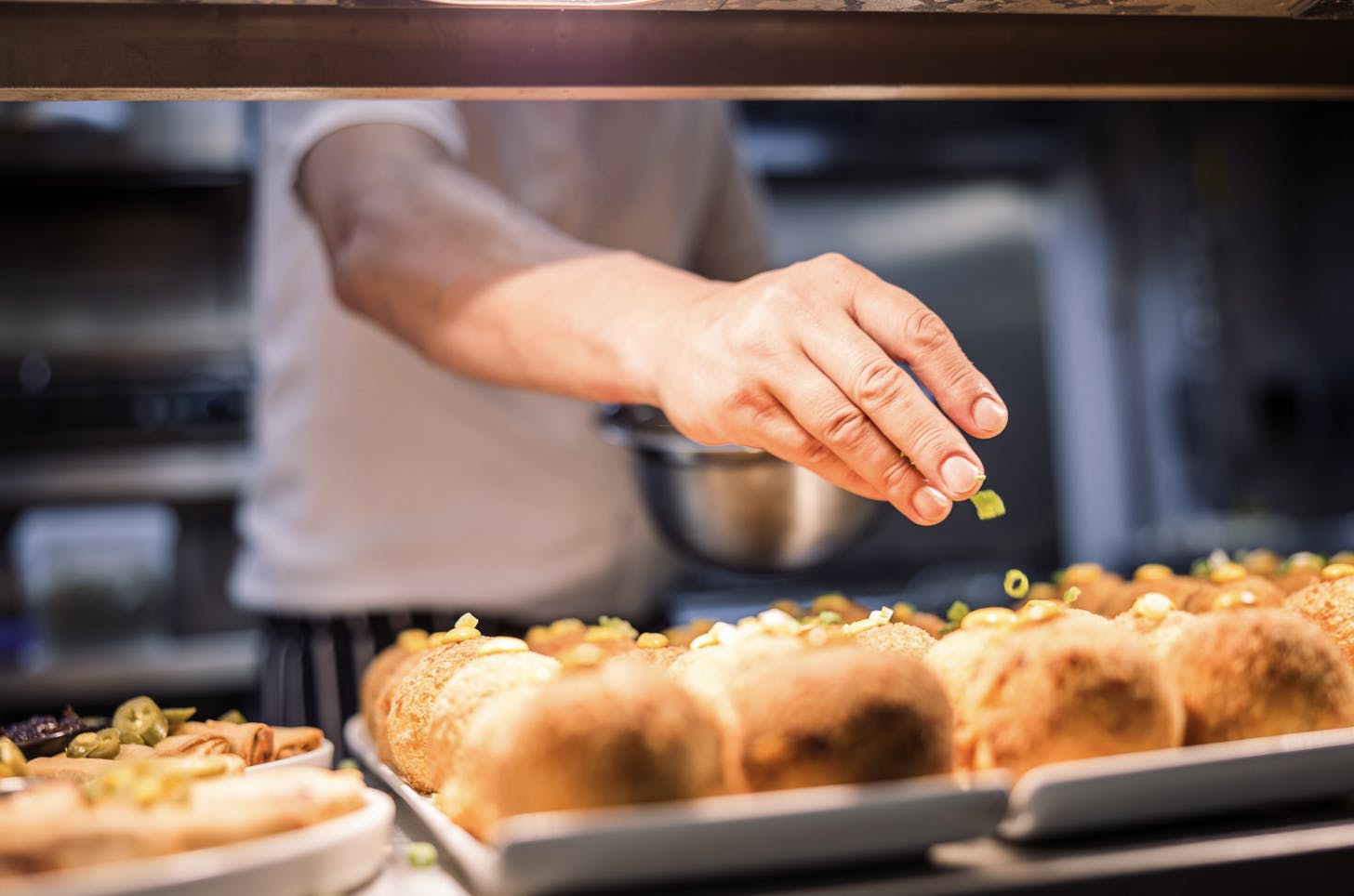 A chef reaches into garnish some large arancini under a hot counter.