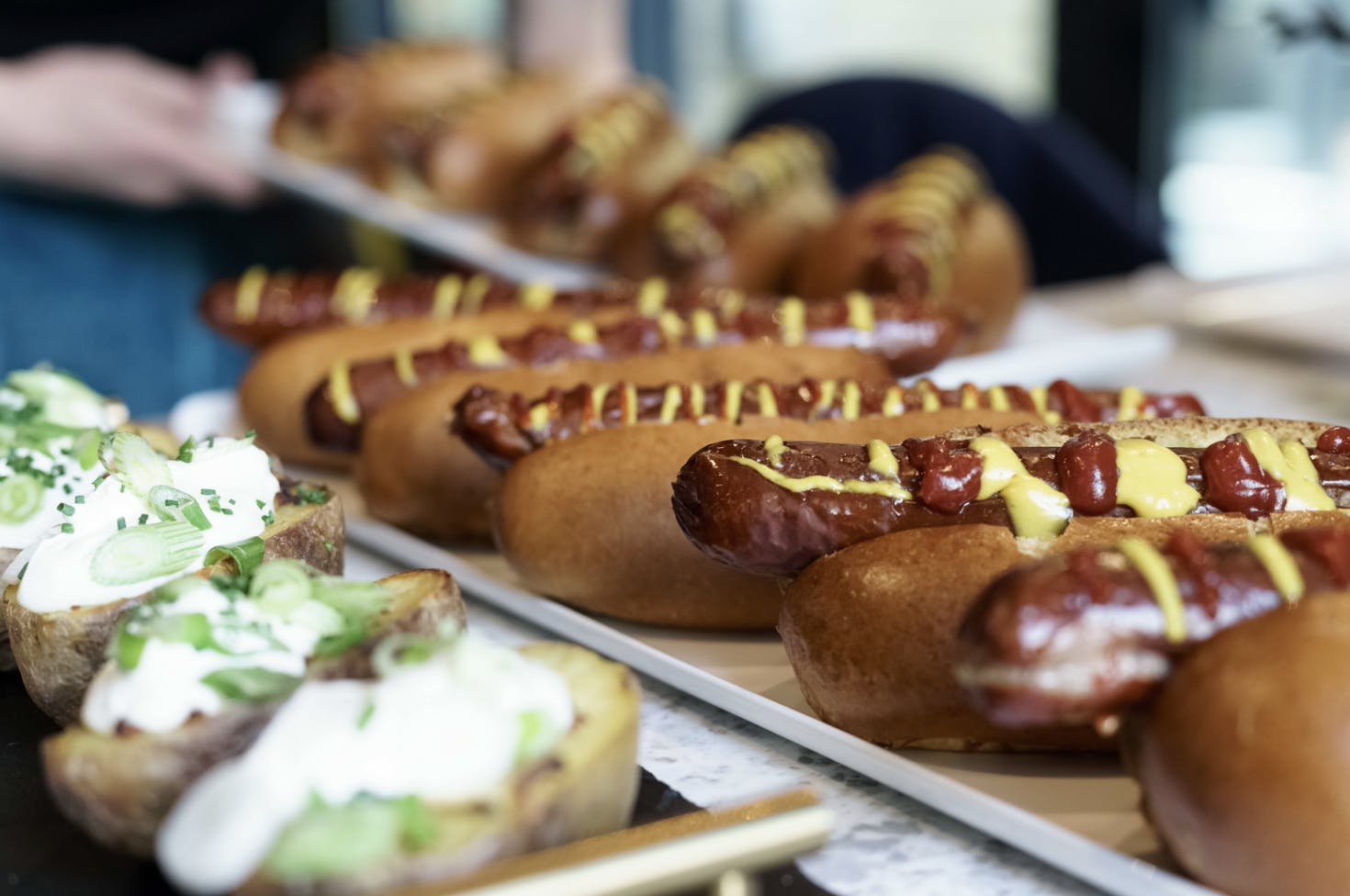 Gourmet hot dogs, stuffed potatoes are part of a buffet spread being laid out.