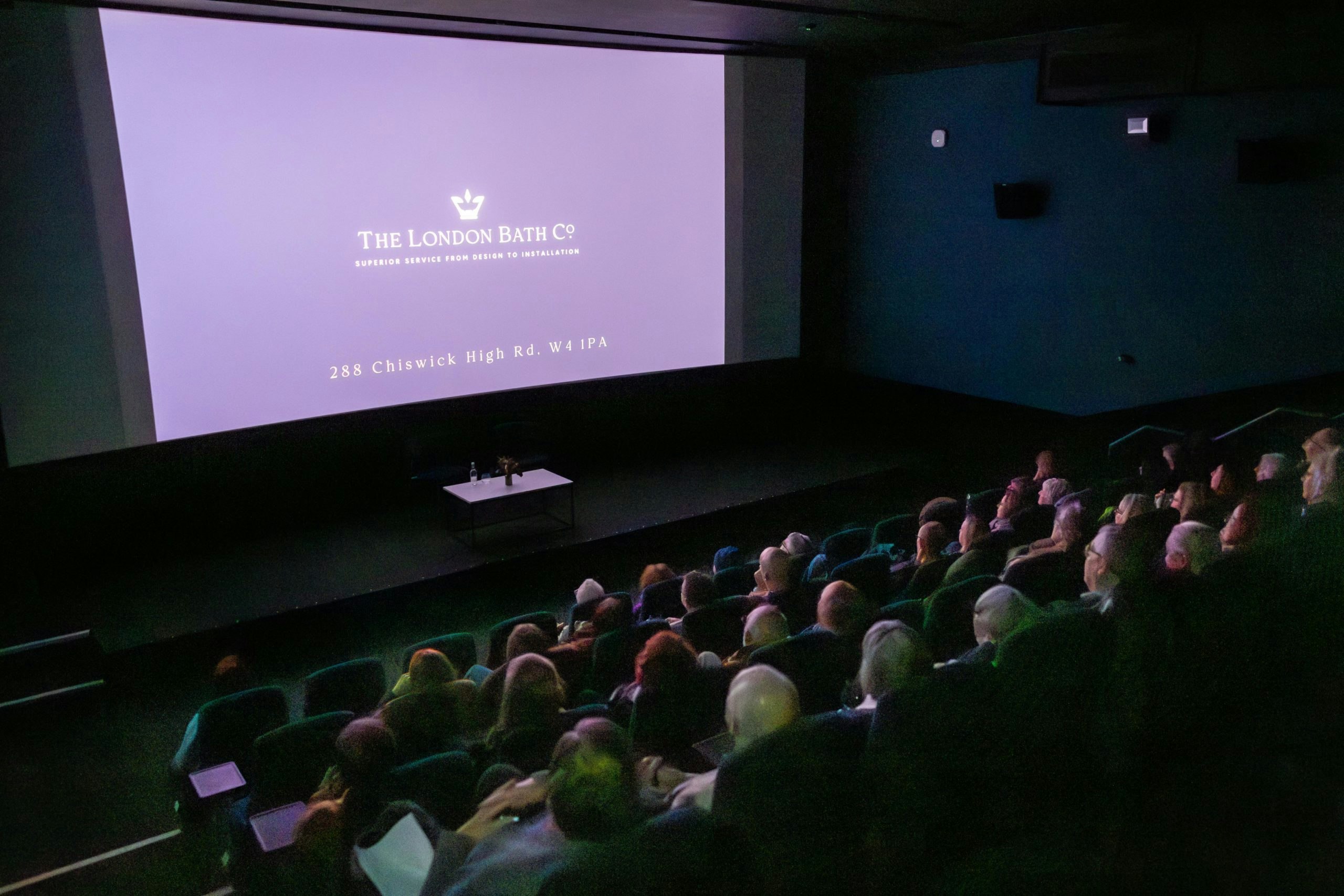 A full audience of people sees the London Bath Co logo on a cinema screen.