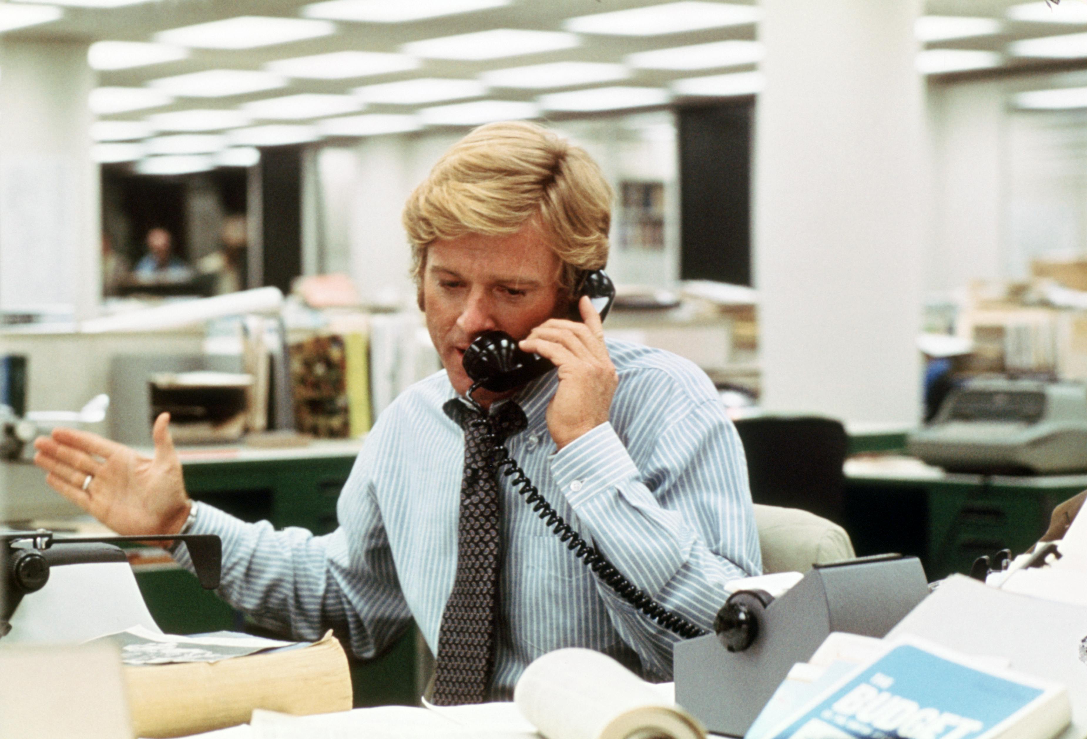 Robert Redford speaking on a telephone inside a busy newsroom
